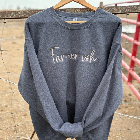 A gray crewneck sweatshirt with the text 'Farmerish' embroidered on the front, displayed on a wooden hanger against a rustic background.