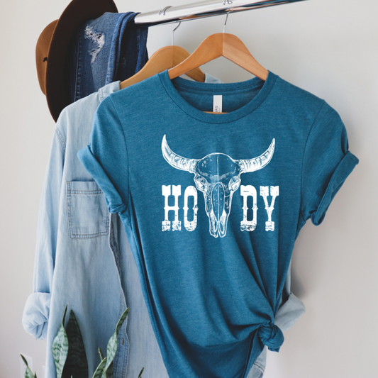 A blue t-shirt with a 'HOWDY' graphic and a bovine skull design displayed on a wooden hanger with denim and a hat on either side.