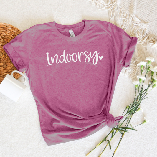 A pink t-shirt with the text 'indoorsy' printed in cursive script, displayed on a cozy white background with a cup of coffee and a decorative bow in the background, accompanied by a bouquet of flowers.