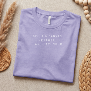 Folded purple t-shirt with 'Bella & Canvas Heather Dark Lavender' text on a beige surface with decorative elements.