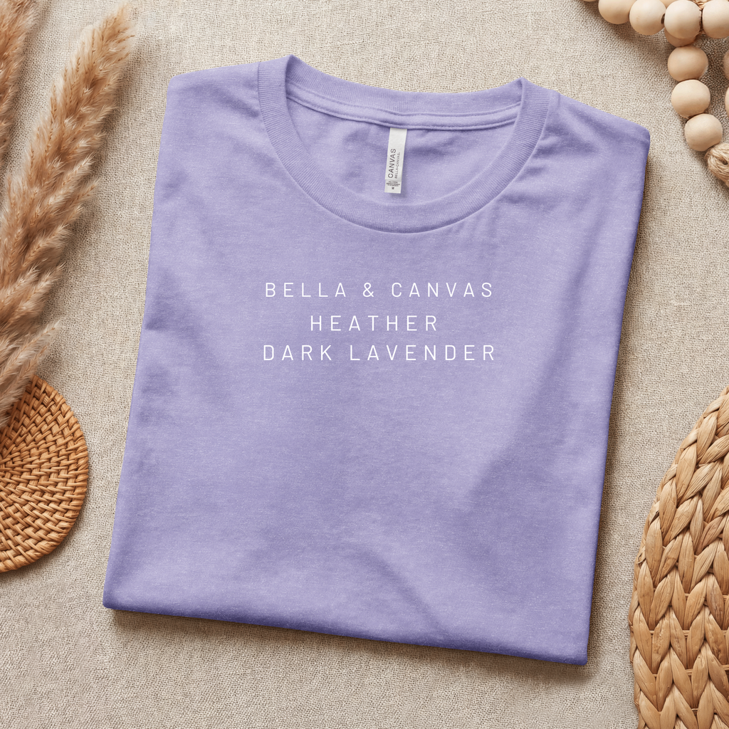 Folded purple t-shirt with 'Bella & Canvas Heather Dark Lavender' text on a beige surface with decorative elements.