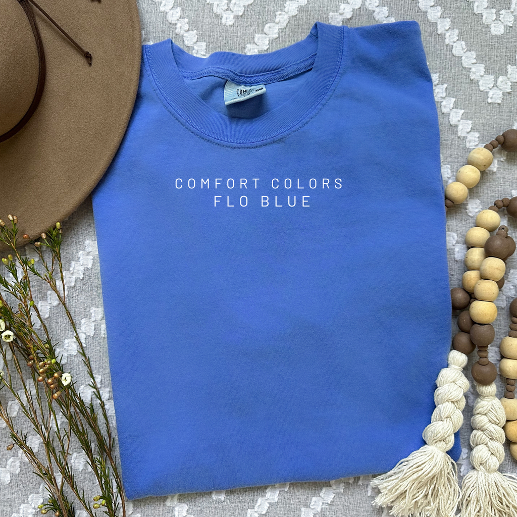 Blue sweatshirt with 'Comfort Colors Flo Blue' text on a textured surface with a hat and beads.