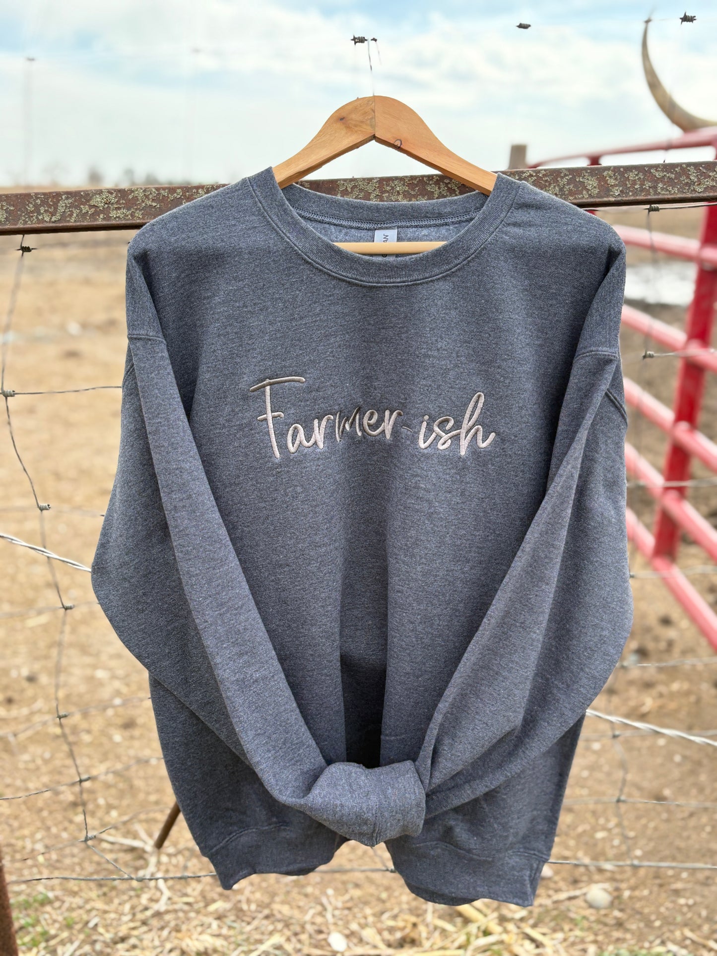 A gray crewneck sweatshirt with the text 'Farmerish' embroidered on the front, displayed on a wooden hanger against a rustic background.