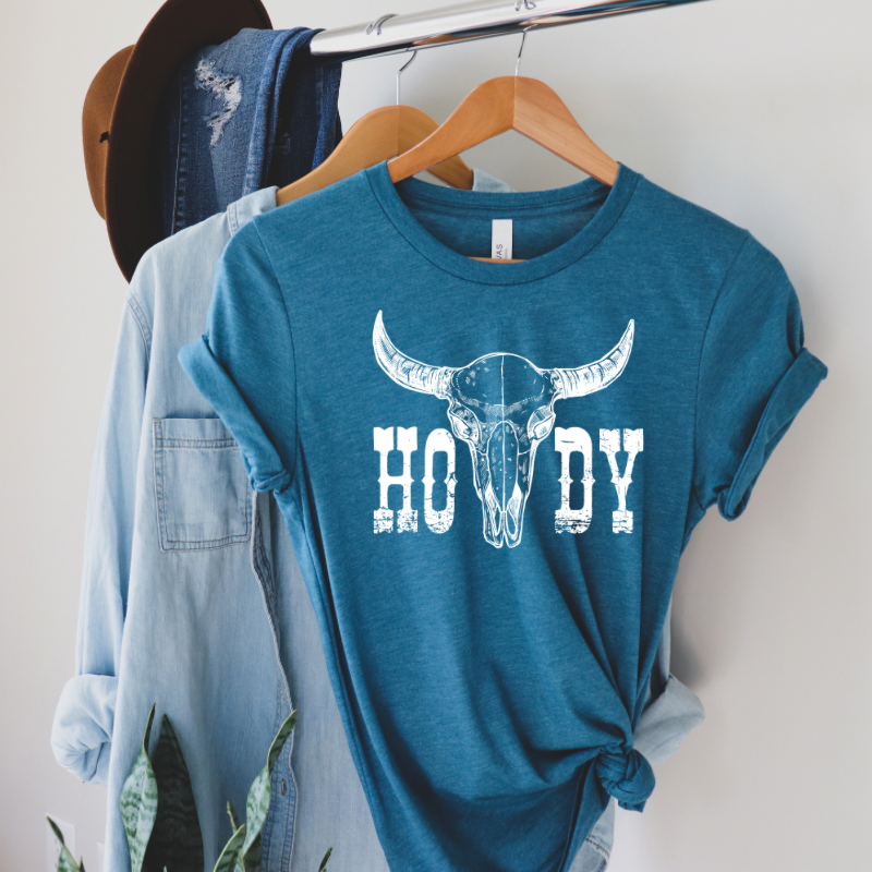 A blue t-shirt with a 'HOWDY' graphic and a bovine skull design displayed on a wooden hanger with denim and a hat on either side.