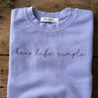 Folded light purple sweatshirt with 'keep life simple' text on a wooden surface
