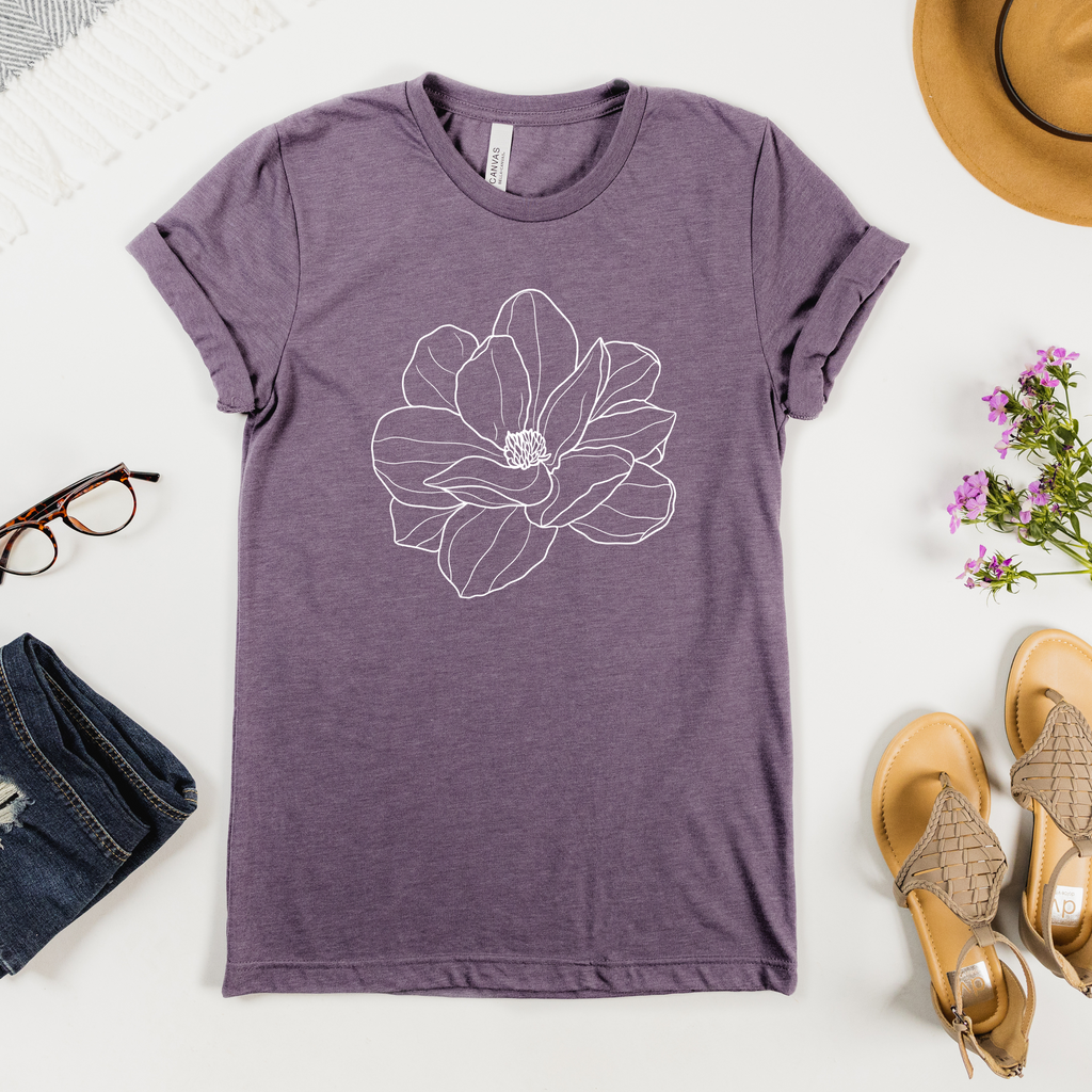 A purple t-shirt with a white magnolia flower design, displayed flat with a pair of jeans, a hat, and prescription glasses on a table.