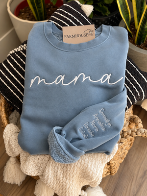 Blue sweatshirt with 'mama' printed on it, displayed on a woven basket with plants in the background.