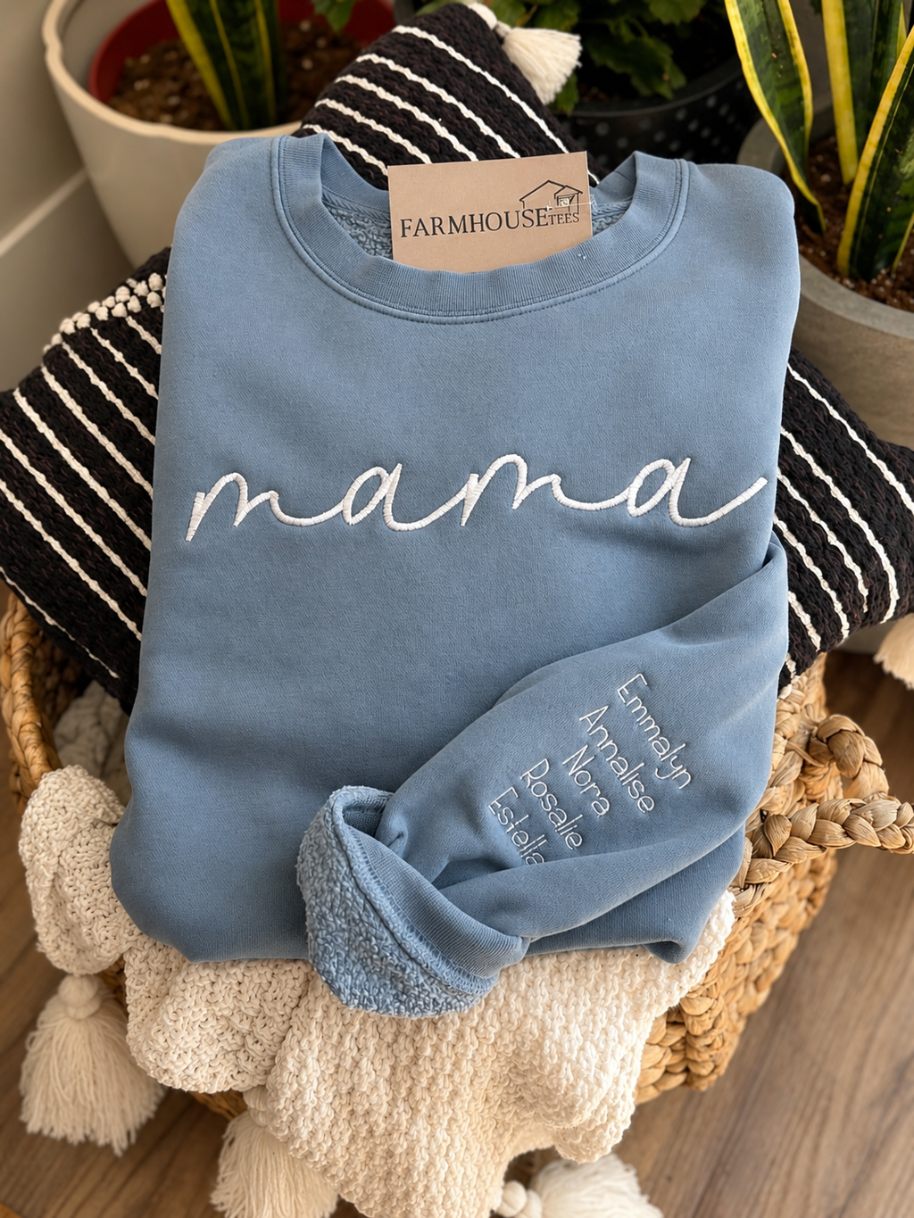 Blue sweatshirt with 'mama' printed on it, displayed on a woven basket with plants in the background.