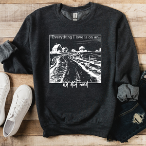 A gray sweatshirt with a graphic print that says 'Everything I love is on an old dirt road' on the front, displayed on a wooden surface with a pair of blue jeans and white sneakers.