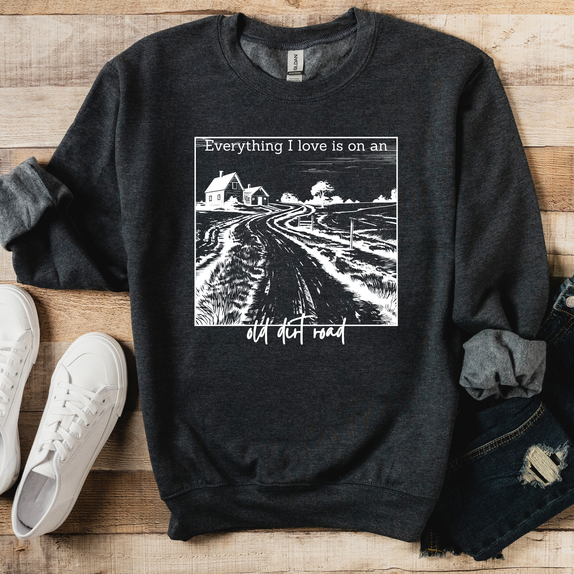 A gray sweatshirt with a graphic print that says 'Everything I love is on an old dirt road' on the front, displayed on a wooden surface with a pair of blue jeans and white sneakers.
