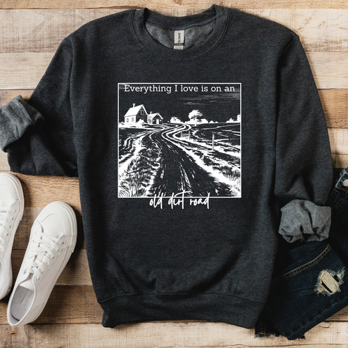 A gray sweatshirt with a graphic print that says 'Everything I love is on an old dirt road' on the front, displayed on a wooden surface with a pair of blue jeans and white sneakers.