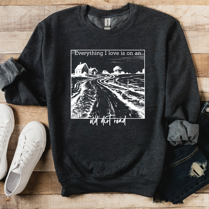 A gray sweatshirt with a graphic print that says 'Everything I love is on an old dirt road' on the front, displayed on a wooden surface with a pair of blue jeans and white sneakers.