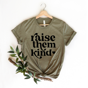 A green t-shirt with the text 'RAISE THEM KIND' printed on the front, displayed on a wooden hanger with a branch and leaves in the background.