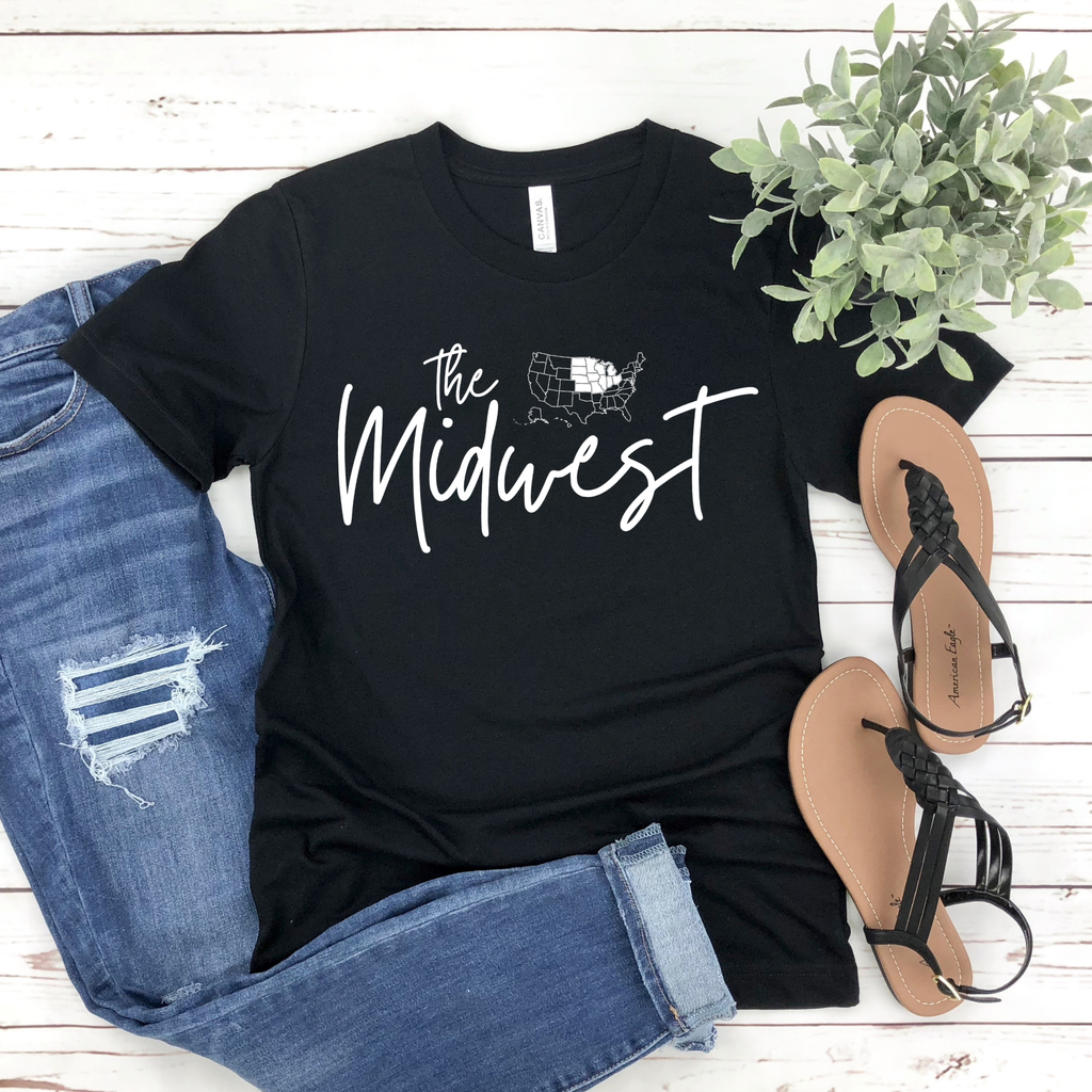 Black t-shirt with 'The Midwest' text, paired with denim shorts and sandals on a wooden surface.