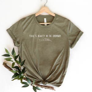 Green t-shirt with 'There is beauty in the ordinary' text on a hanger with a branch and leaves.
