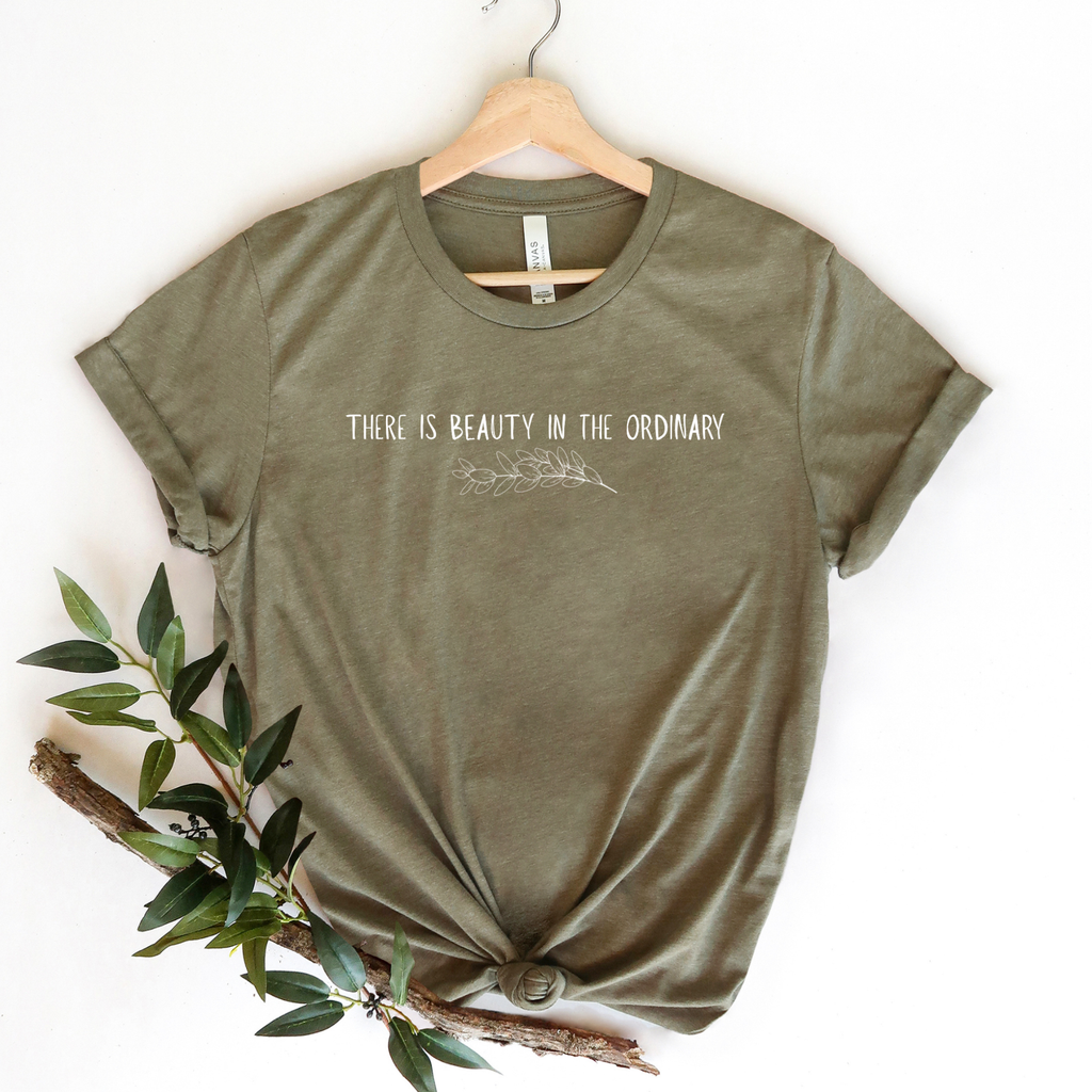 Green t-shirt with 'There is beauty in the ordinary' text on a hanger with a branch and leaves.
