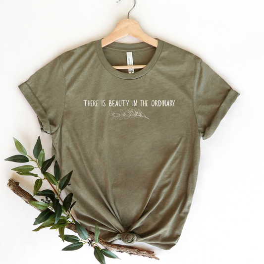 Green t-shirt with 'There is beauty in the ordinary' text on a hanger with a branch and leaves.