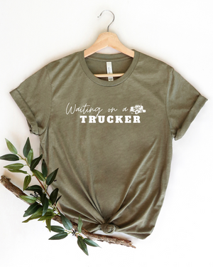 Olive green t-shirt with 'Waiting on a Trucker' text on a hanger with a branch and leaves.