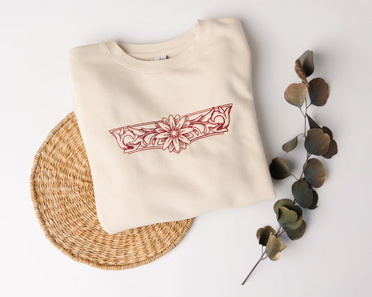 Beige sweatshirt with red floral design on a white background with a woven mat and dried leaves.
