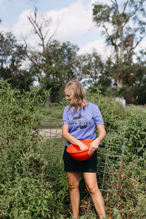 Woman in a garden holding an orange bucket, wearing a blue shirt with text 'support your local farm wives'.
