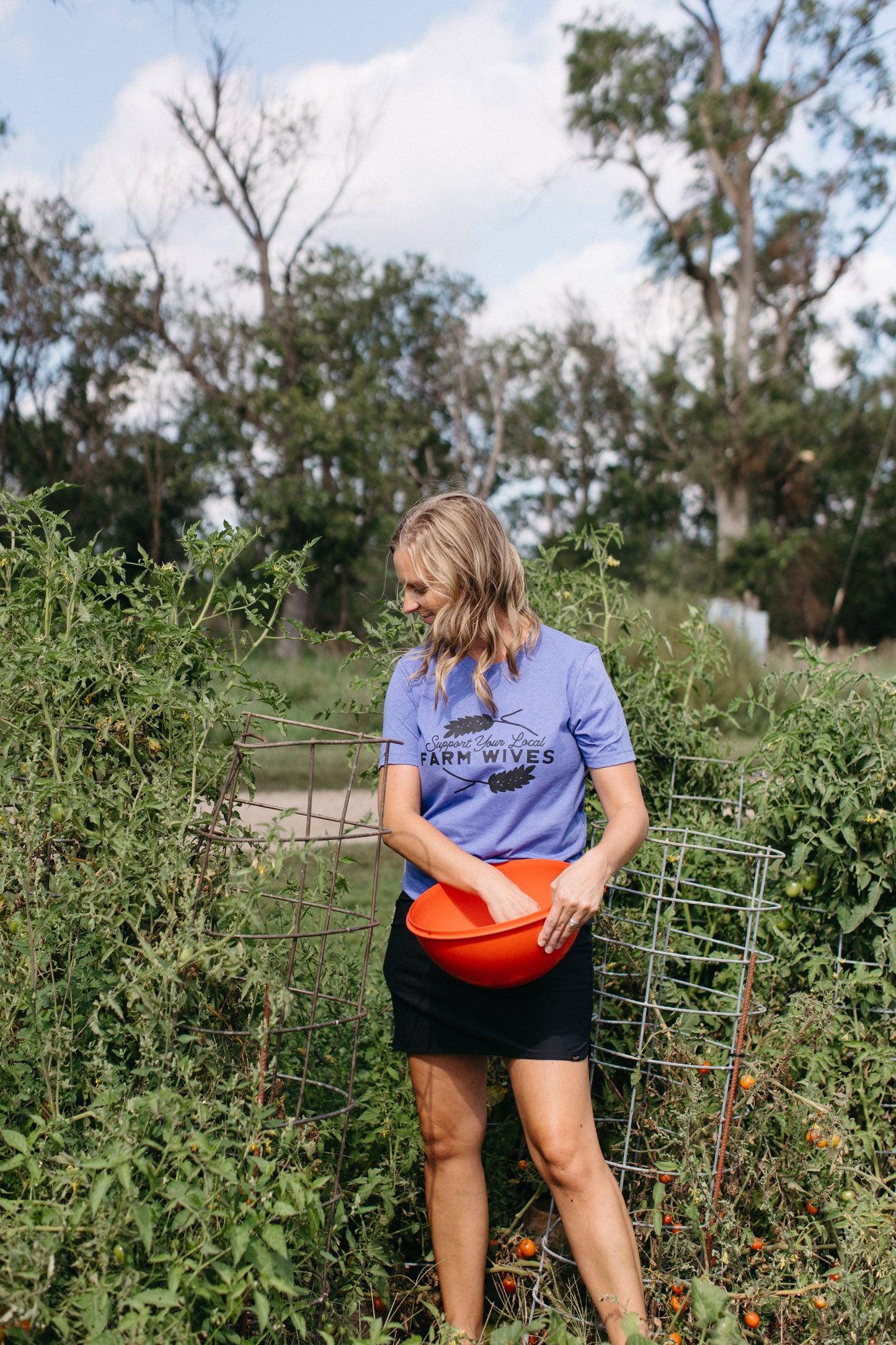 Woman in a garden holding an orange bucket, wearing a blue shirt with text 'support your local farm wives'.