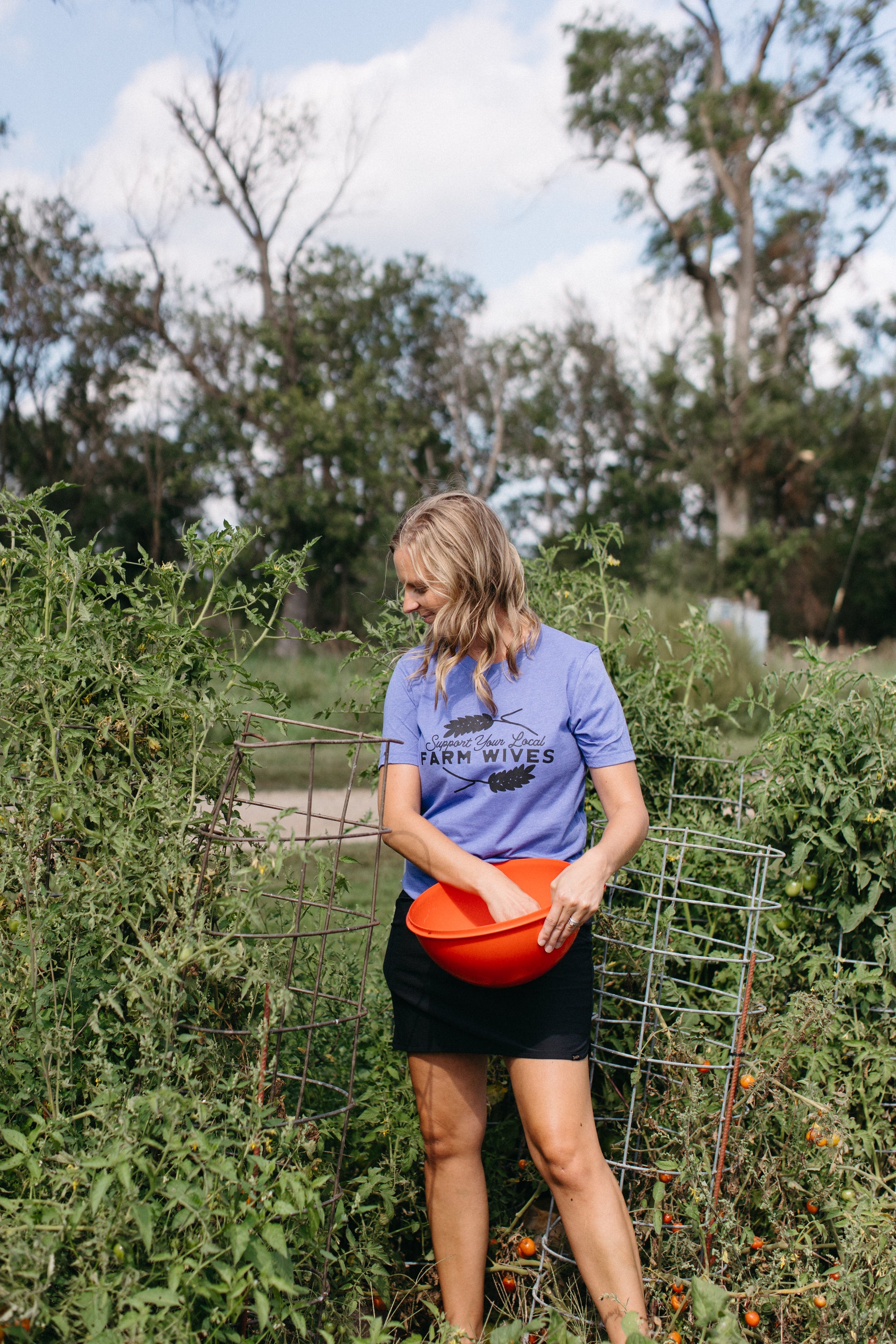 Woman in a garden holding an orange bucket, wearing a blue shirt with text 'support your local farm wives'.