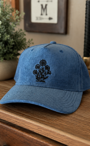 Two corduroy baseball caps shown side by side in a split-screen layout. The left cap is medium blue with a black embroidered floral design, resting on a wooden surface.