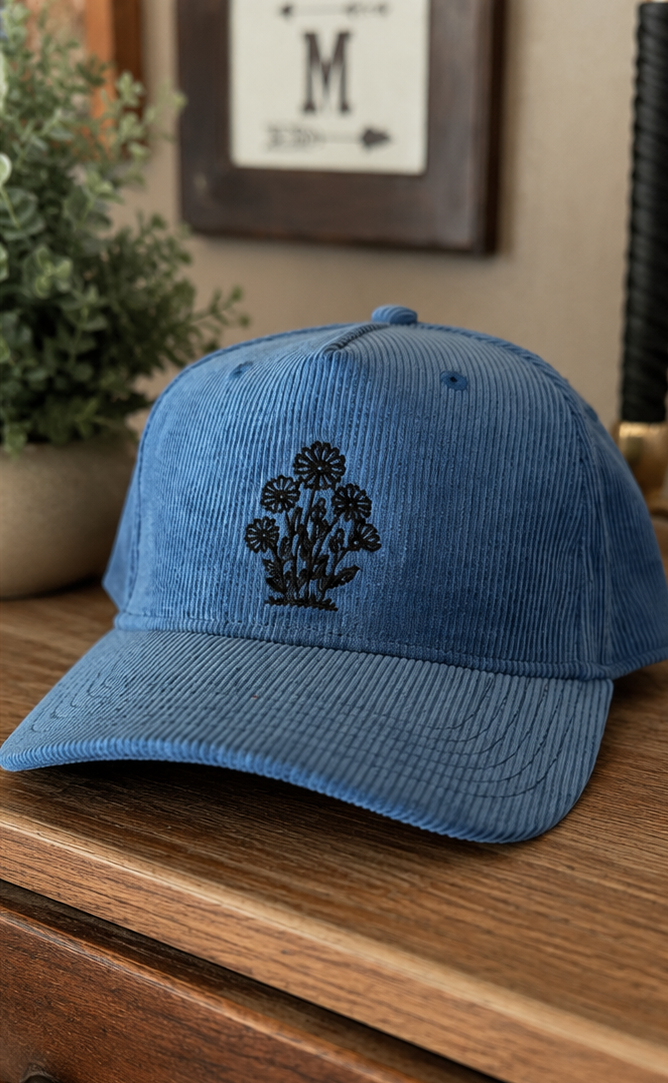 Two corduroy baseball caps shown side by side in a split-screen layout. The left cap is medium blue with a black embroidered floral design, resting on a wooden surface.