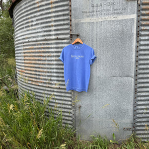 a blue tee with text 'farm mom'  hanging on a metal surface with grass and trees in the background