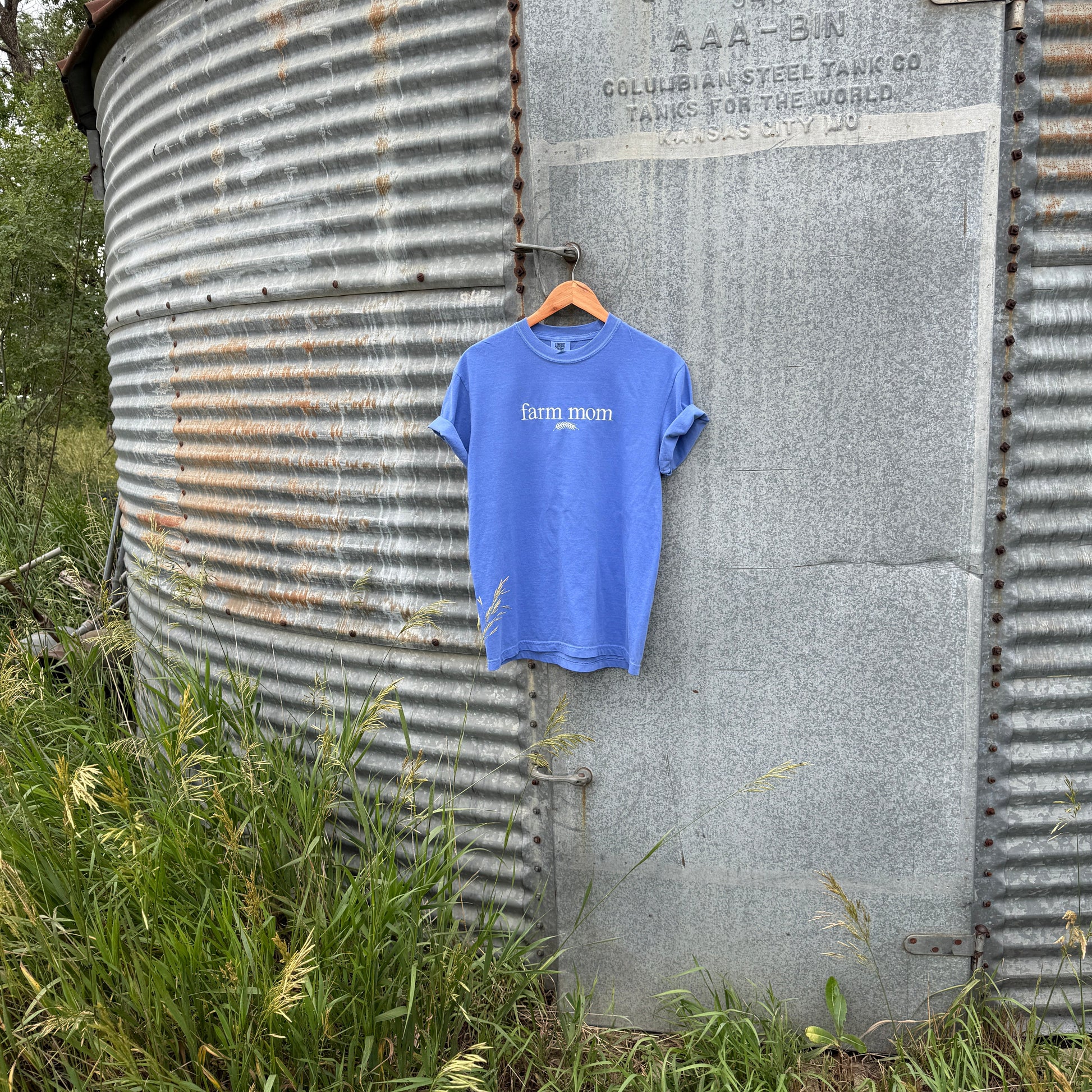 a blue tee with text 'farm mom'  hanging on a metal surface with grass and trees in the background
