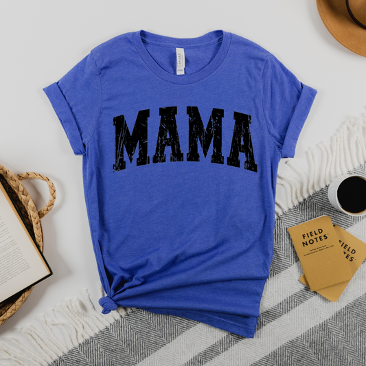 A blue t-shirt with the word 'MAMA' printed in black text on the front, displayed on a flat surface with a hat, books, and a cardboard sign in the background.