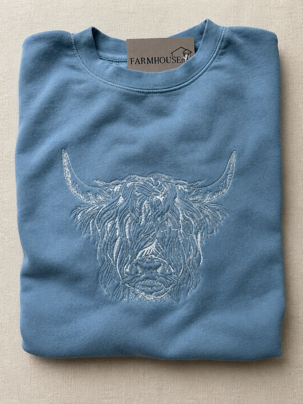 Folded blue sweatshirt with a highland cow design and 'Farmhouse' label on a beige background