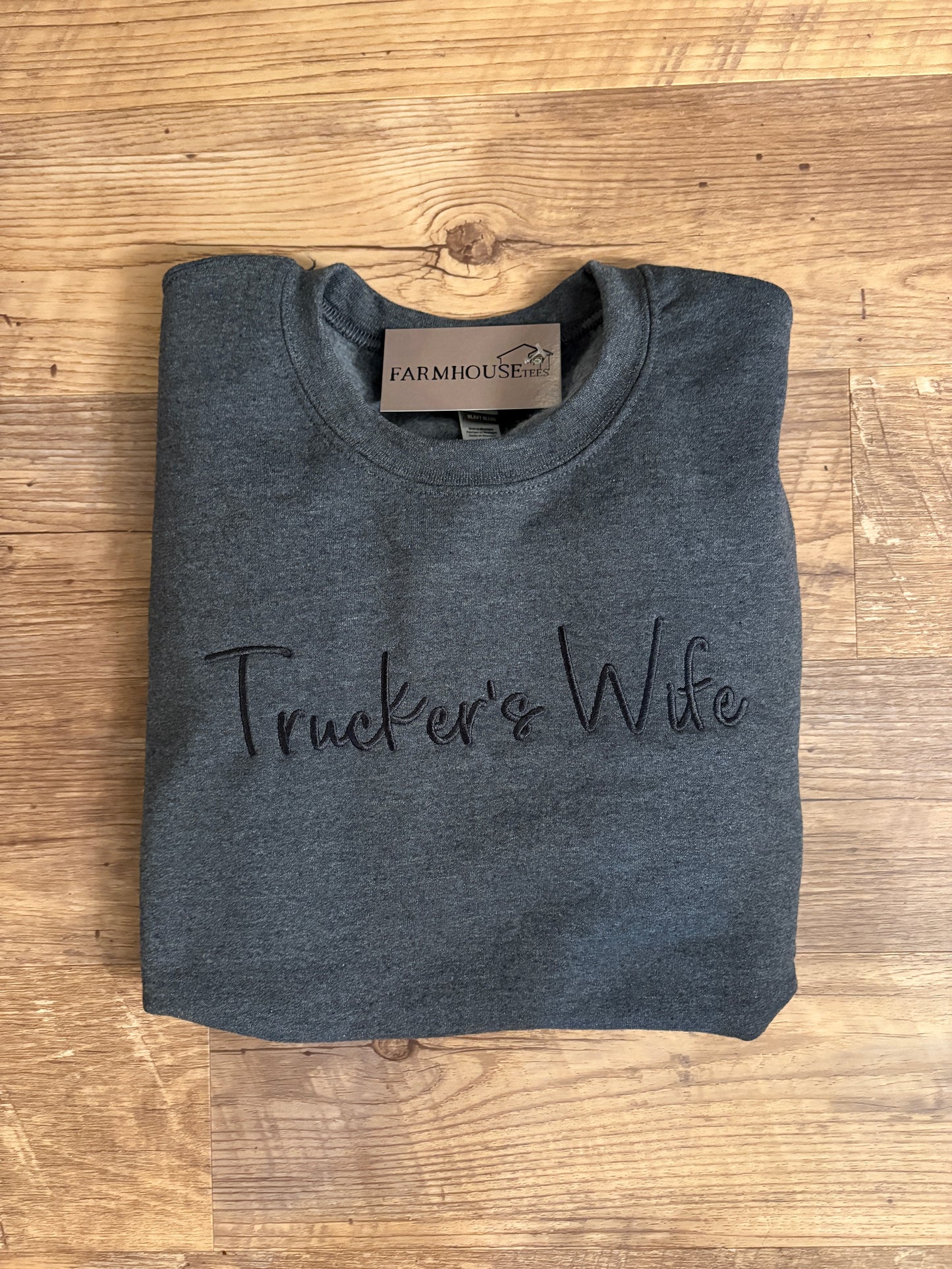 Folded gray t-shirt with 'Trucker's Wife' text on a wooden surface