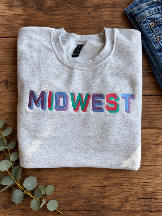 A light heather gray sweatshirt with the word 'MIDWEST' printed across the chest in colorful, textured block letters. The sweatshirt is styled in a flat-lay arrangement on a rustic wooden table, accompanied by blue jeans, a brown cowboy hat, and dried wheat stalks.