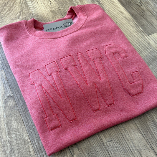 Folded pink sweatshirt with 'NWC' embroidery on a wooden surface