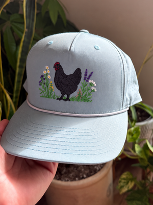 A light blue baseball cap held by a hand, featuring an embroidered design of a black chicken standing among green and purple flowers on the front panel. The background shows potted plants in a home setting.