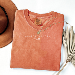 Folded orange shirt with 'Comfort Colors' label, accompanied by a hat and dried plant on a white background