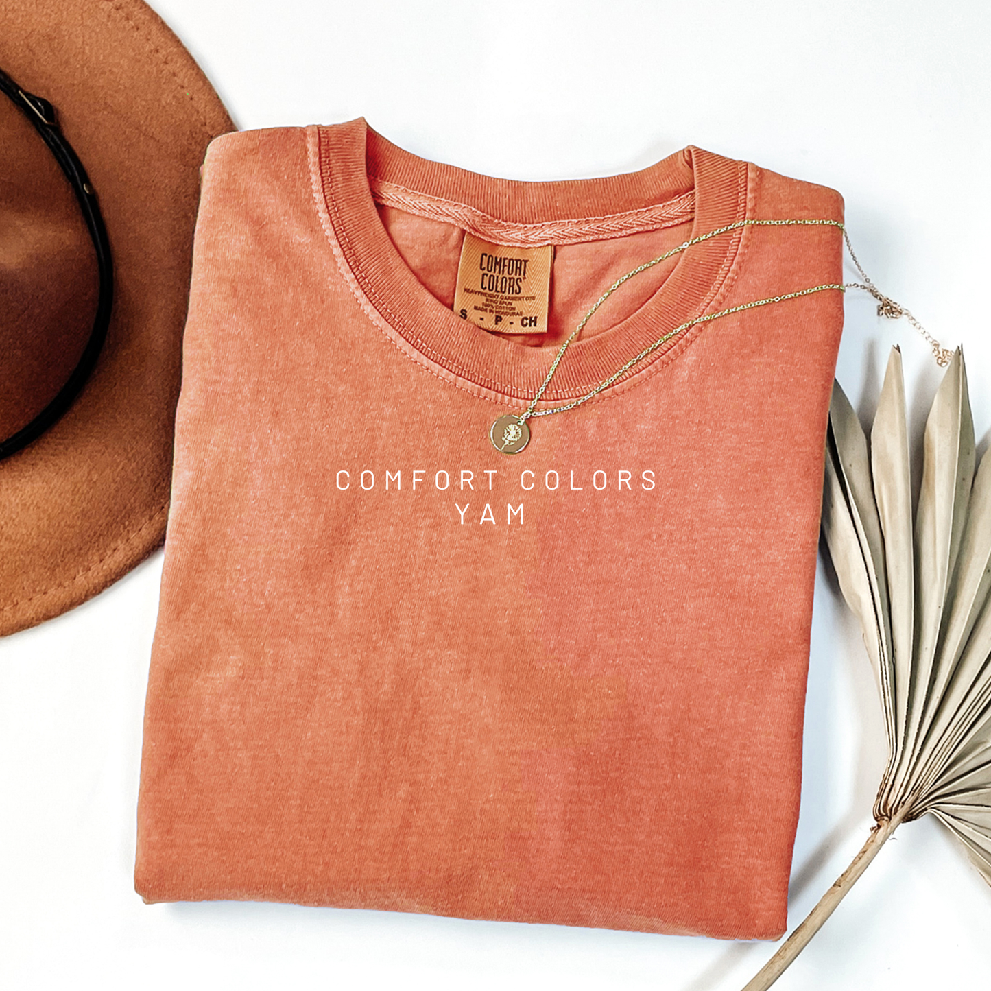 Folded orange shirt with 'Comfort Colors' label, accompanied by a hat and dried plant on a white background