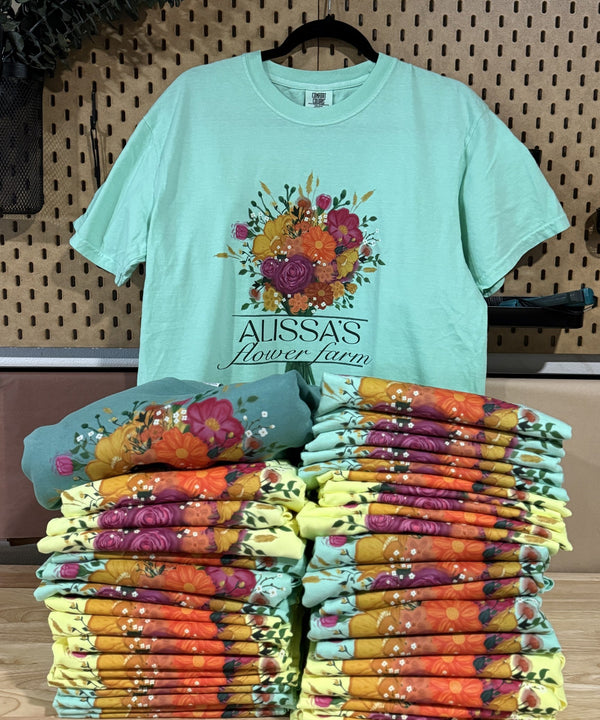 A mint green graphic t-shirt with a colorful floral print and the text 'ALISSA'S Flower Farm' hanging on a black hanger against a pegboard wall. In front of the shirt, stacks of folded t-shirts in various colorful floral patterns are neatly arranged on a wooden table.