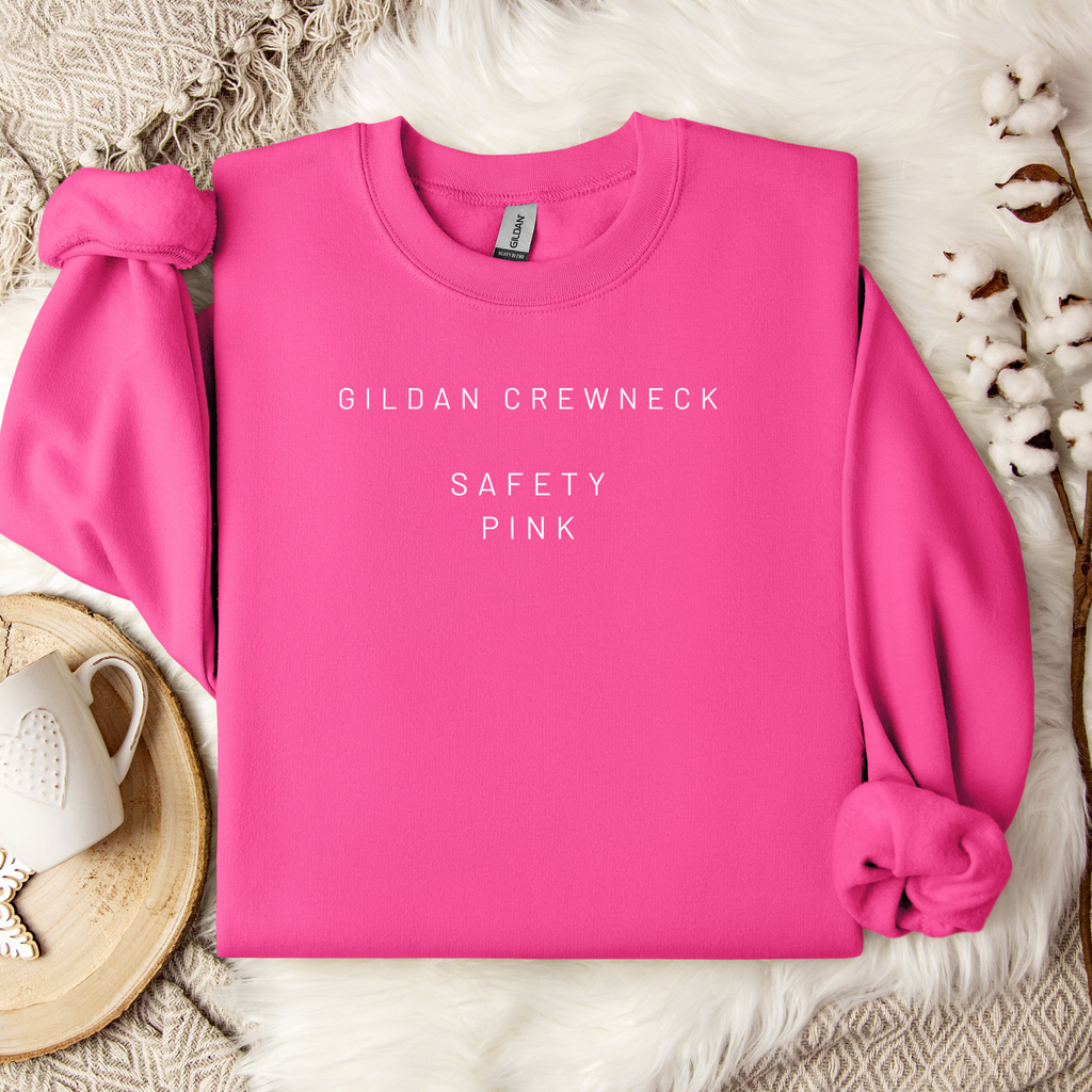 Pink Gildan crewneck sweatshirt on a textured surface with a cup and decorative elements.