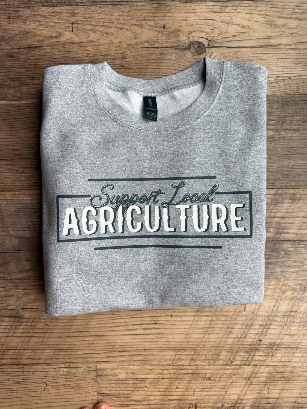 Folded gray t-shirt with 'support your local Agriculture' text on a wooden surface
