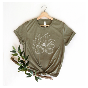 A green Bella Canvas T-shirt with a white magnolia flower design, displayed on a hanger with a plant decoration to the side.