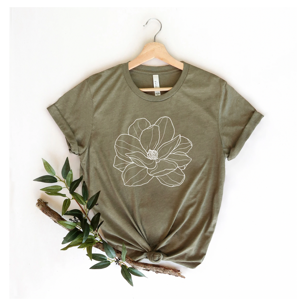 A green Bella Canvas T-shirt with a white magnolia flower design, displayed on a hanger with a plant decoration to the side.