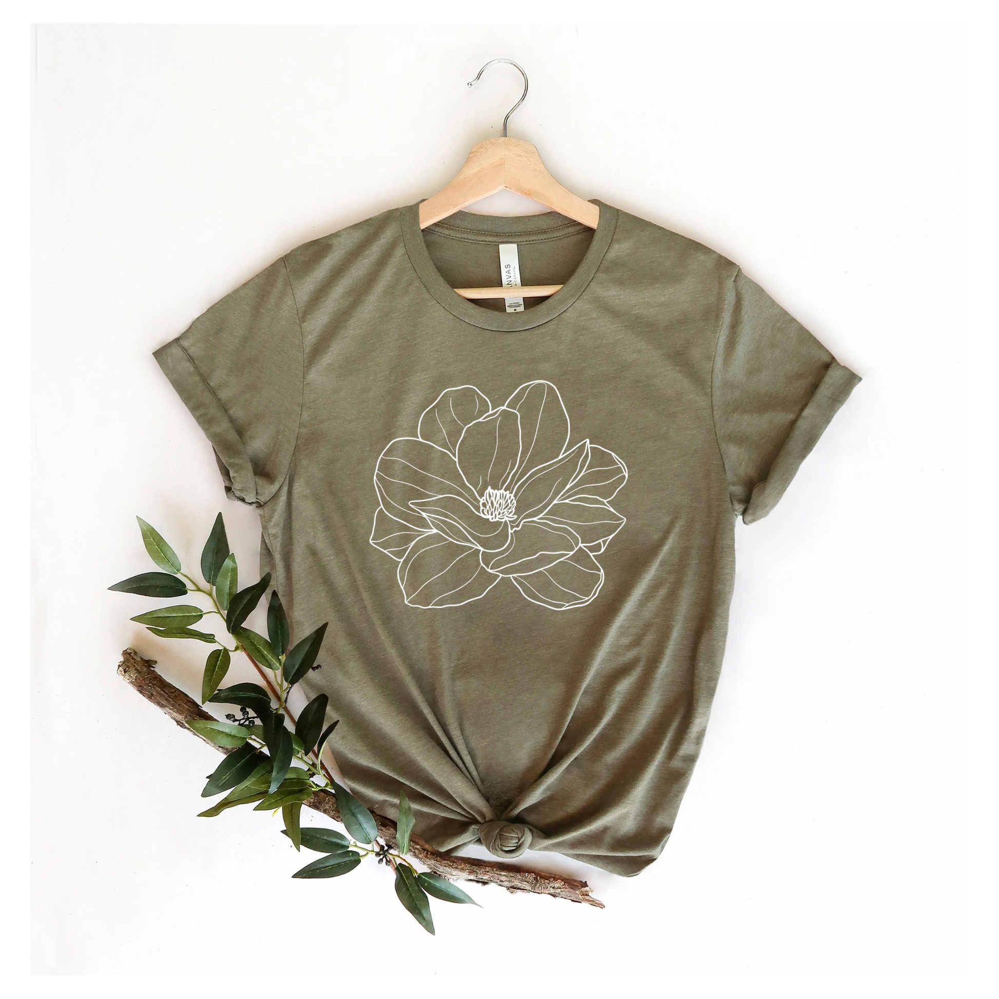 A green Bella Canvas T-shirt with a white magnolia flower design, displayed on a hanger with a plant decoration to the side.