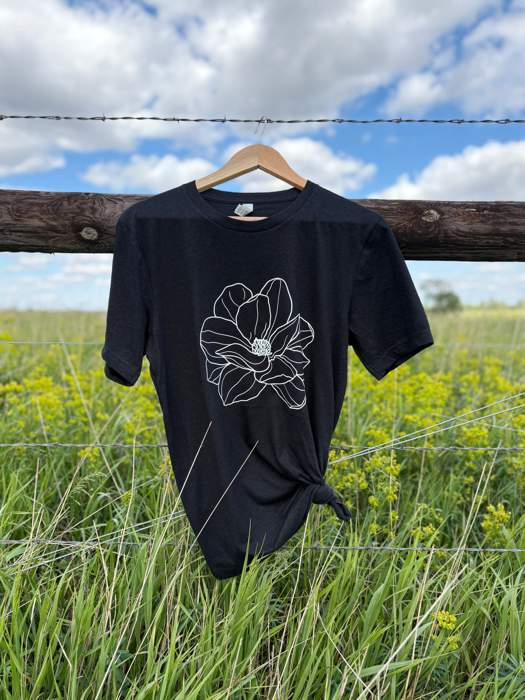 Black t-shirt with a white floral design on a wooden hanger against a natural background of grass and sky.