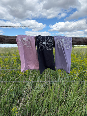 Three fabric shirts with floral print hanging on a wooden post against a blue sky and green grass background.