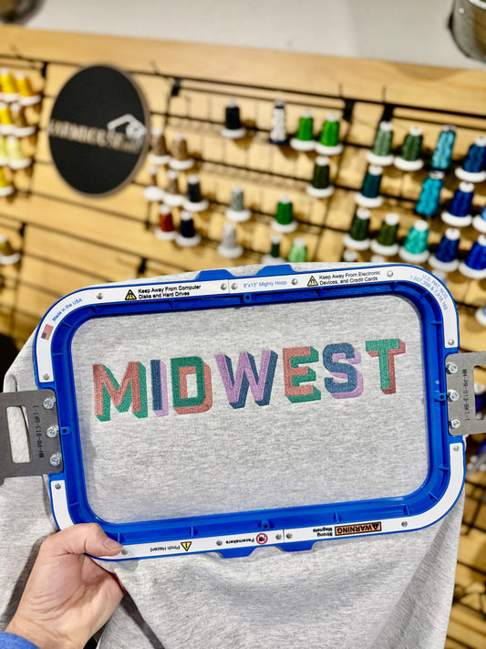 A light heather gray sweatshirt with the word 'MIDWEST' printed across the chest in colorful, textured block letters. The sweatshirt is styled in a flat-lay arrangement on a rustic wooden table, accompanied by blue jeans, a brown cowboy hat, and dried wheat stalks.