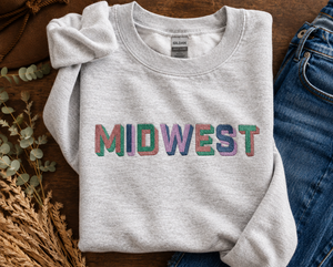 A light heather gray sweatshirt with the word 'MIDWEST' printed across the chest in colorful, textured block letters. The sweatshirt is styled in a flat-lay arrangement on a rustic wooden table, accompanied by blue jeans, a brown cowboy hat, and dried wheat stalks.
