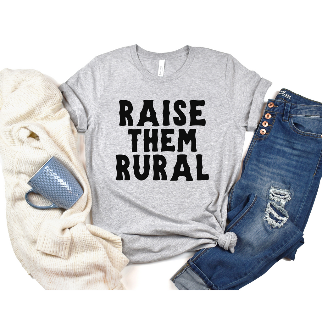 A gray t-shirt with the text 'RAISE THEM RURAL' printed on the front, accompanied by a pair of blue jeans and a textured gray sweater, placed against a white background.