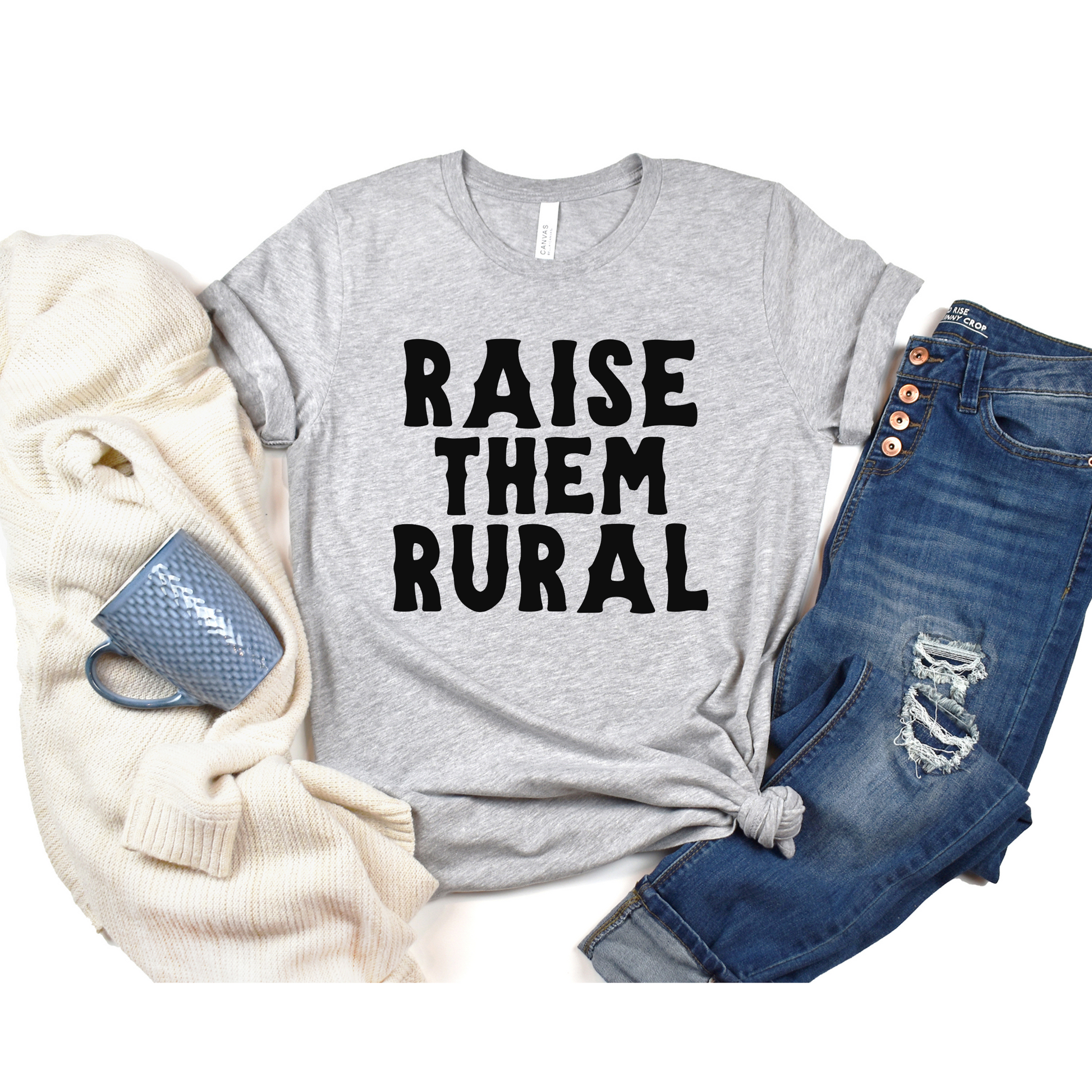 A gray t-shirt with the text 'RAISE THEM RURAL' printed on the front, accompanied by a pair of blue jeans and a textured gray sweater, placed against a white background.